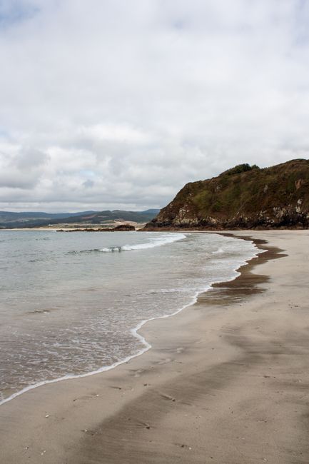 Peaceful beach view with waves lapping on a sandy shore under cloudy skies.