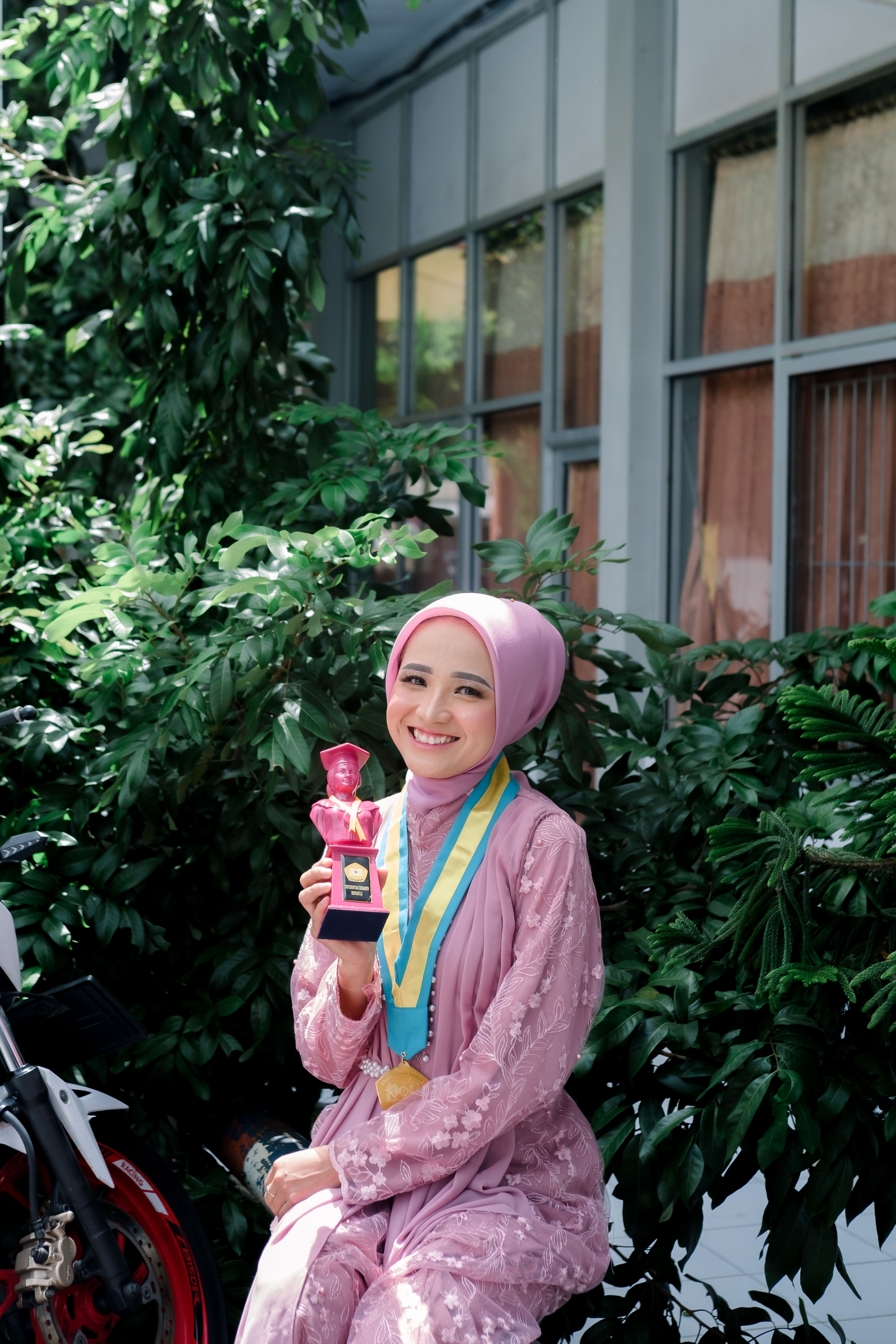 A woman in hijab joyfully poses with her award and medal, conveying academic success.