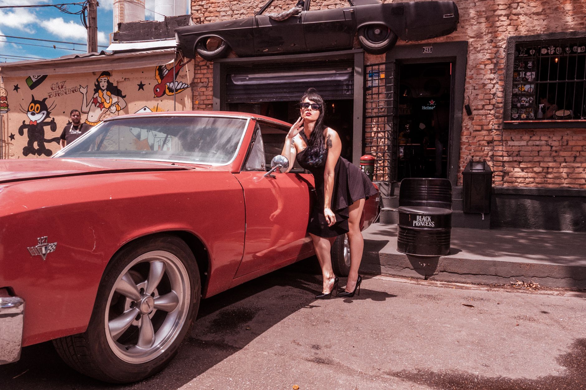 Stylish woman in black dress poses with a vintage red car in an urban setting.