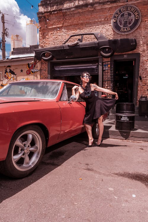 Woman in Black Dress Standing Beside a Red Vintage Car