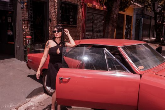 A fashionable woman in sunglasses beside a classic red car on an urban street, exuding retro charm.