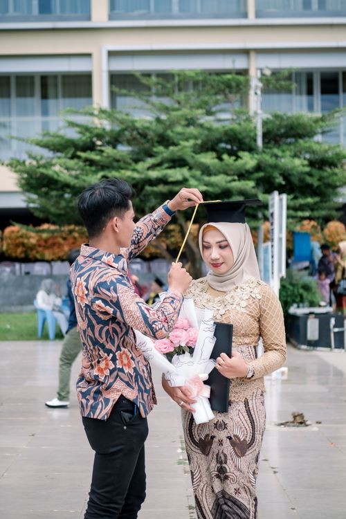 Free A man places an academic hat on a graduate holding a bouquet, celebrating outdoors. Stock Photo