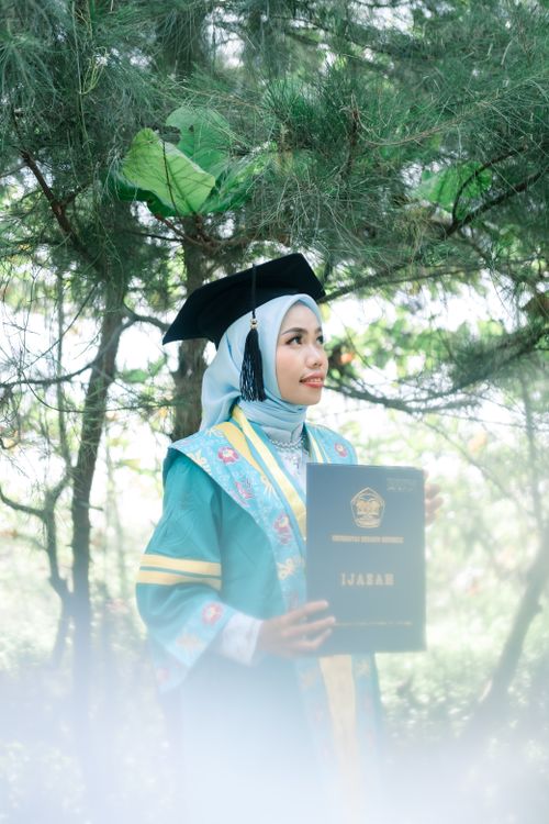 Free A smiling woman in a hijab and gown holds her diploma in a serene outdoor setting. Stock Photo