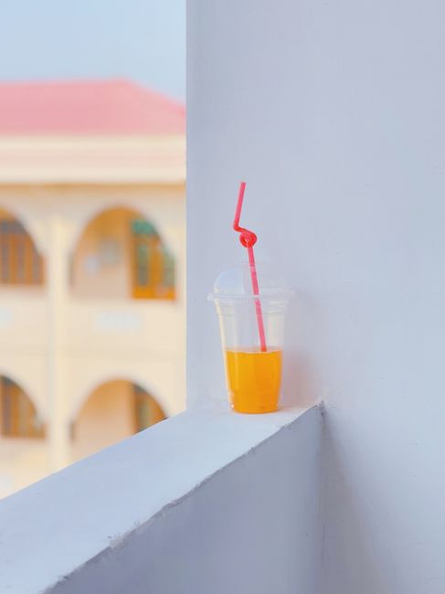A disposable plastic cup with orange juice and pink straw on ledge against blurred background.