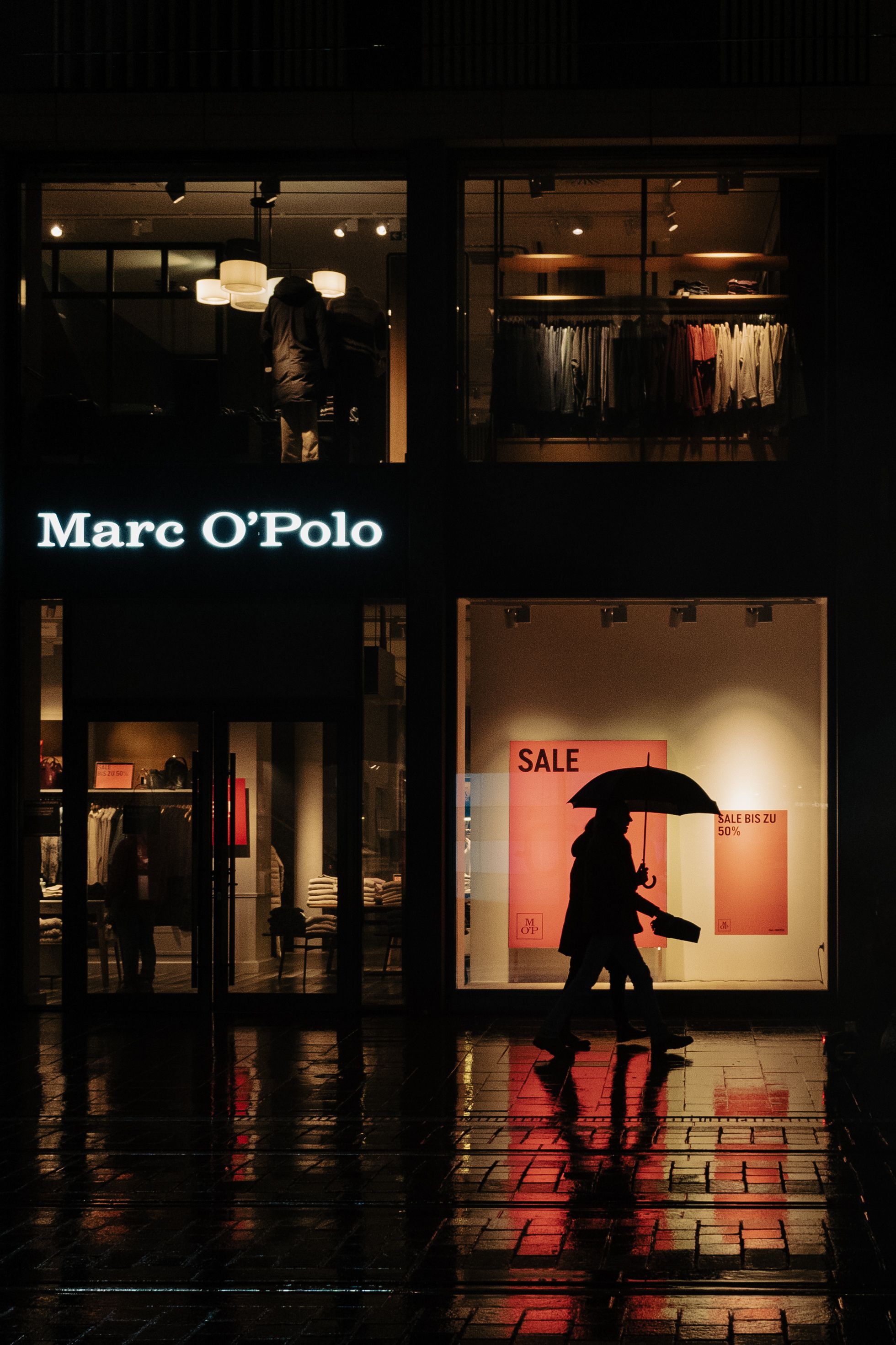 A silhouette of a couple walking with an umbrella in front of a clothing store with a sale sign at night.