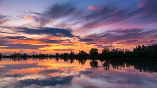 Free Vivid sunset skies reflected on a calm lake with silhouetted trees, creating a mesmerizing scenery. Stock Photo