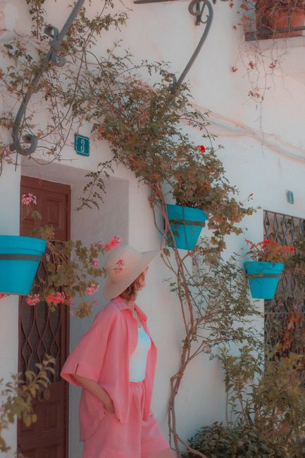 Fashionable woman in pink attire stands by a quaint facade with blue flowerpots