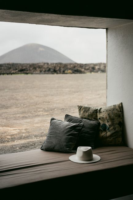 A serene view from a window seat with cushions and hat overlooking a vast desert landscape and distant hill.