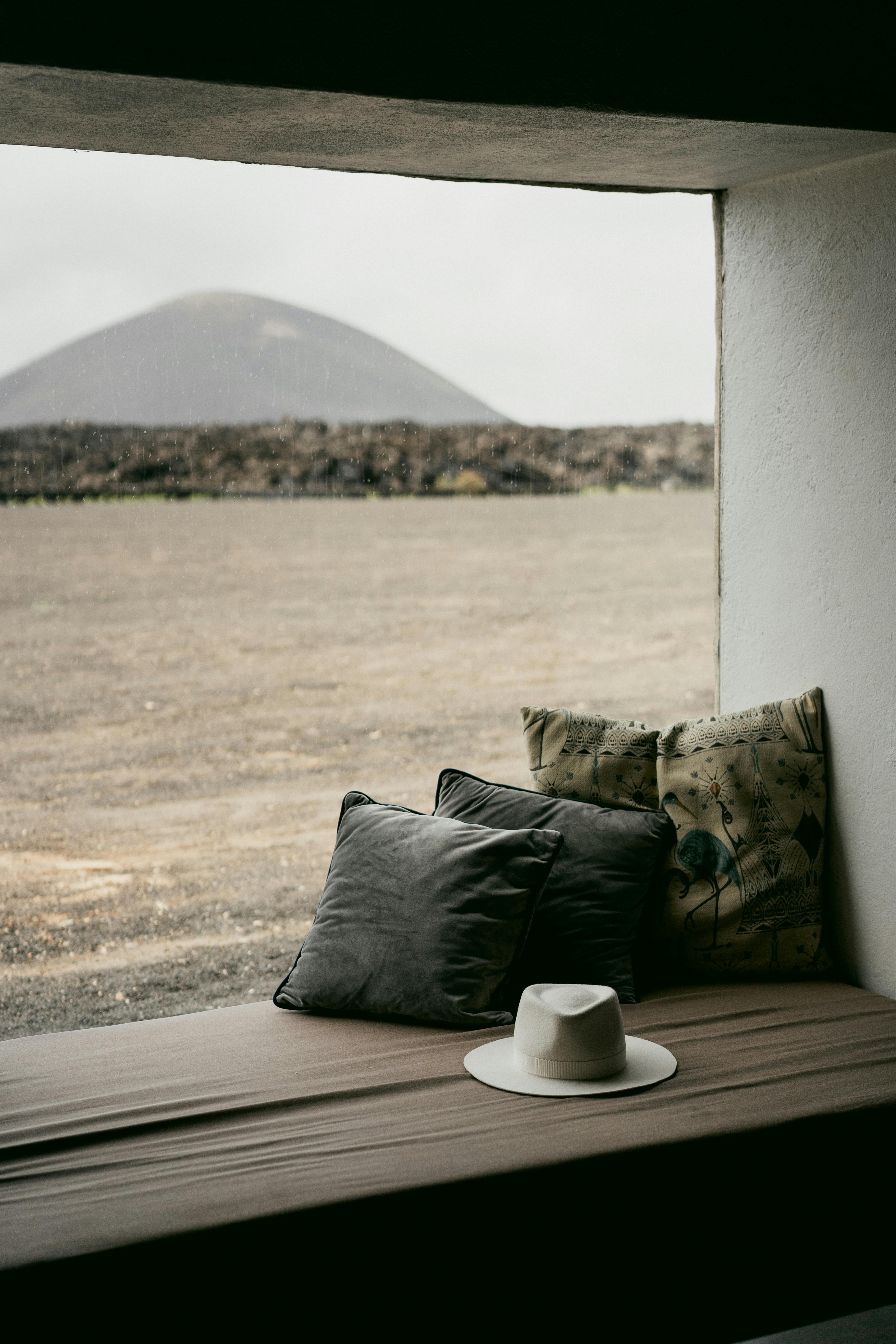 Free A serene view from a window seat with cushions and hat overlooking a vast desert landscape and distant hill. Stock Photo