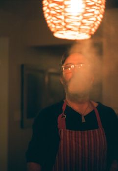 Indoor portrait of a man in an apron standing under warm light, surrounded by smoke.