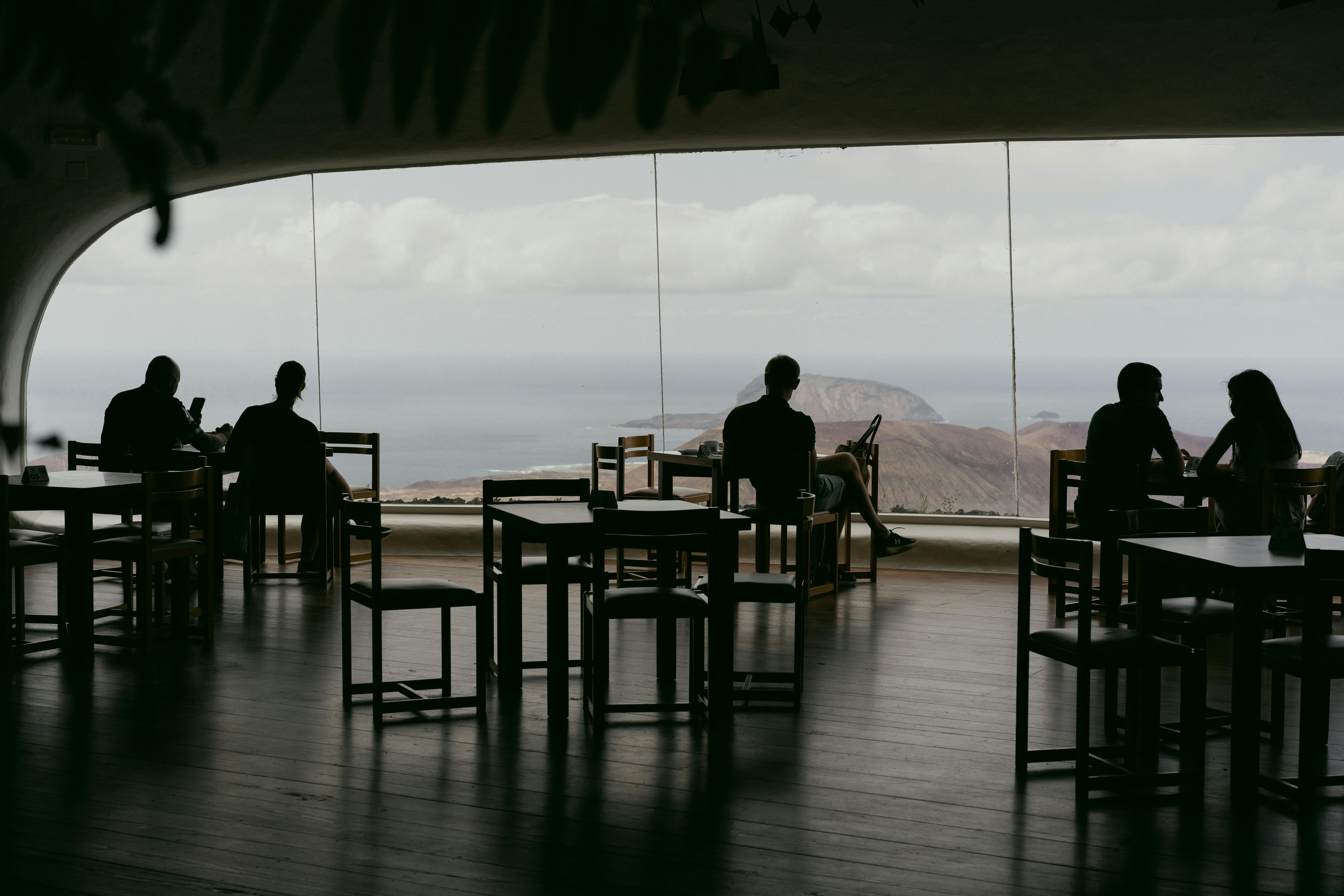 Silhouetted diners enjoy a serene ocean view from a stylish restaurant with large windows.