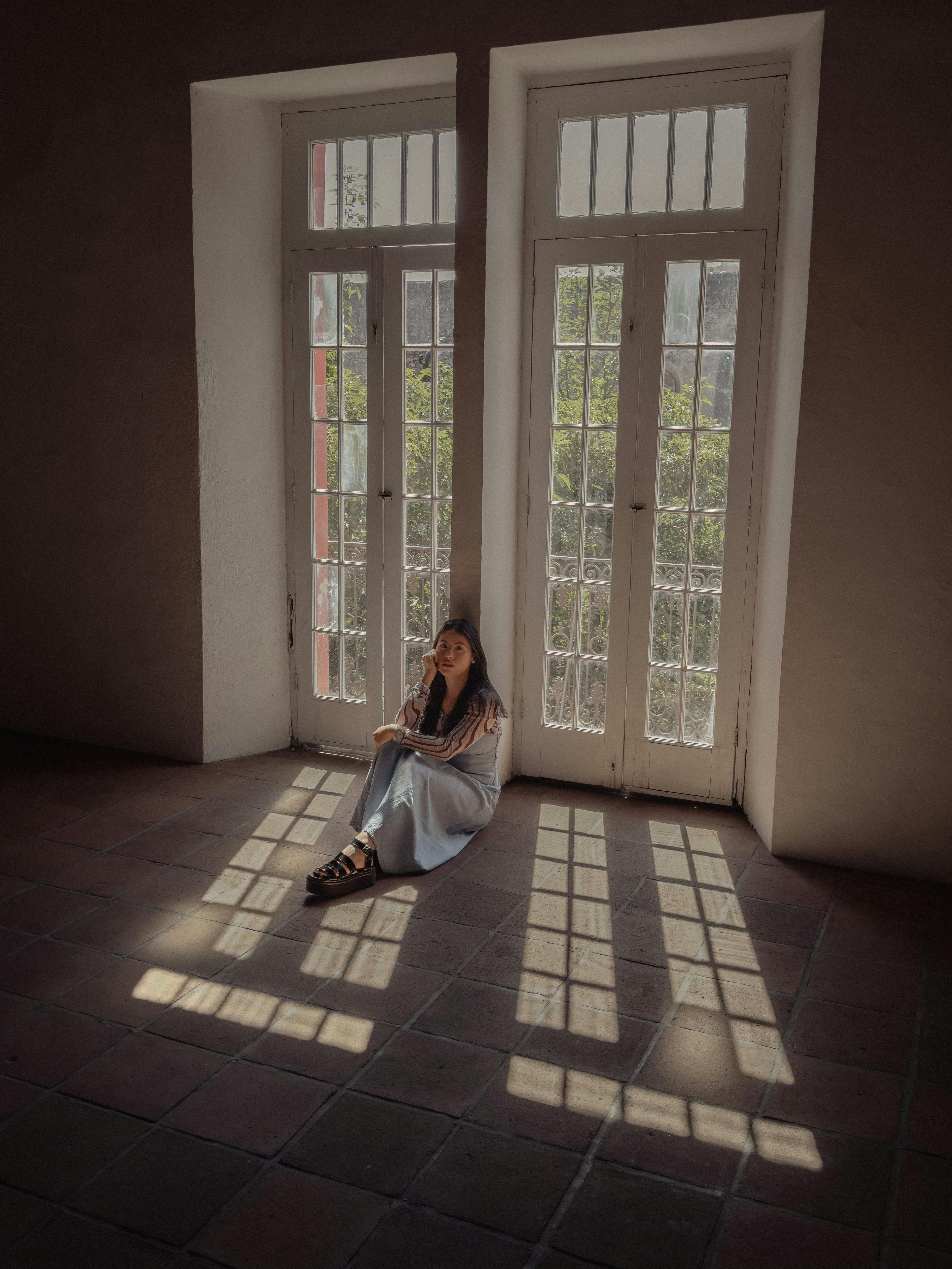 A woman sitting peacefully by tall windows in Ciudad de México, casting geometric shadows on the floor.