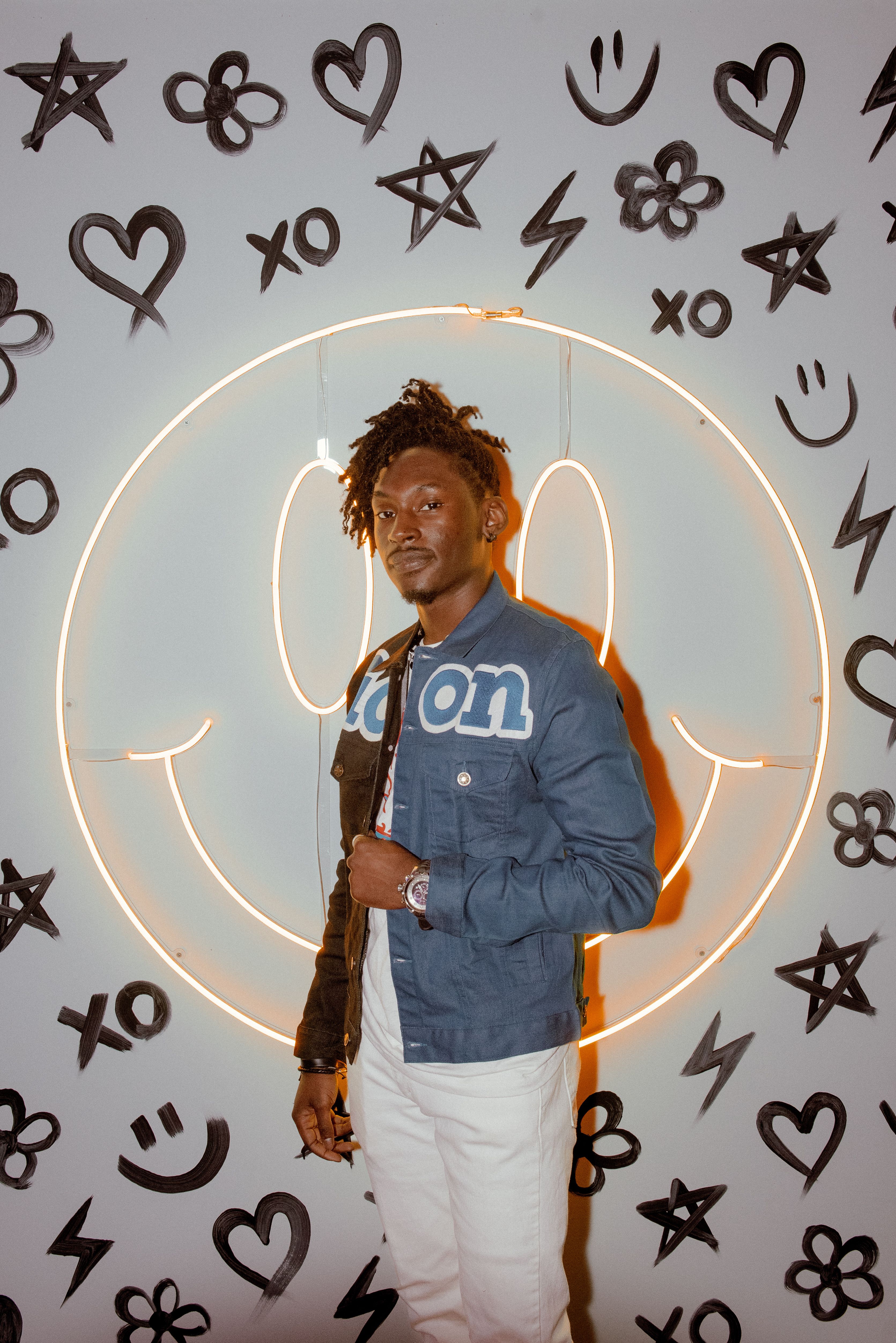 Stylish man in jacket poses against a playful neon smiley wall backdrop.