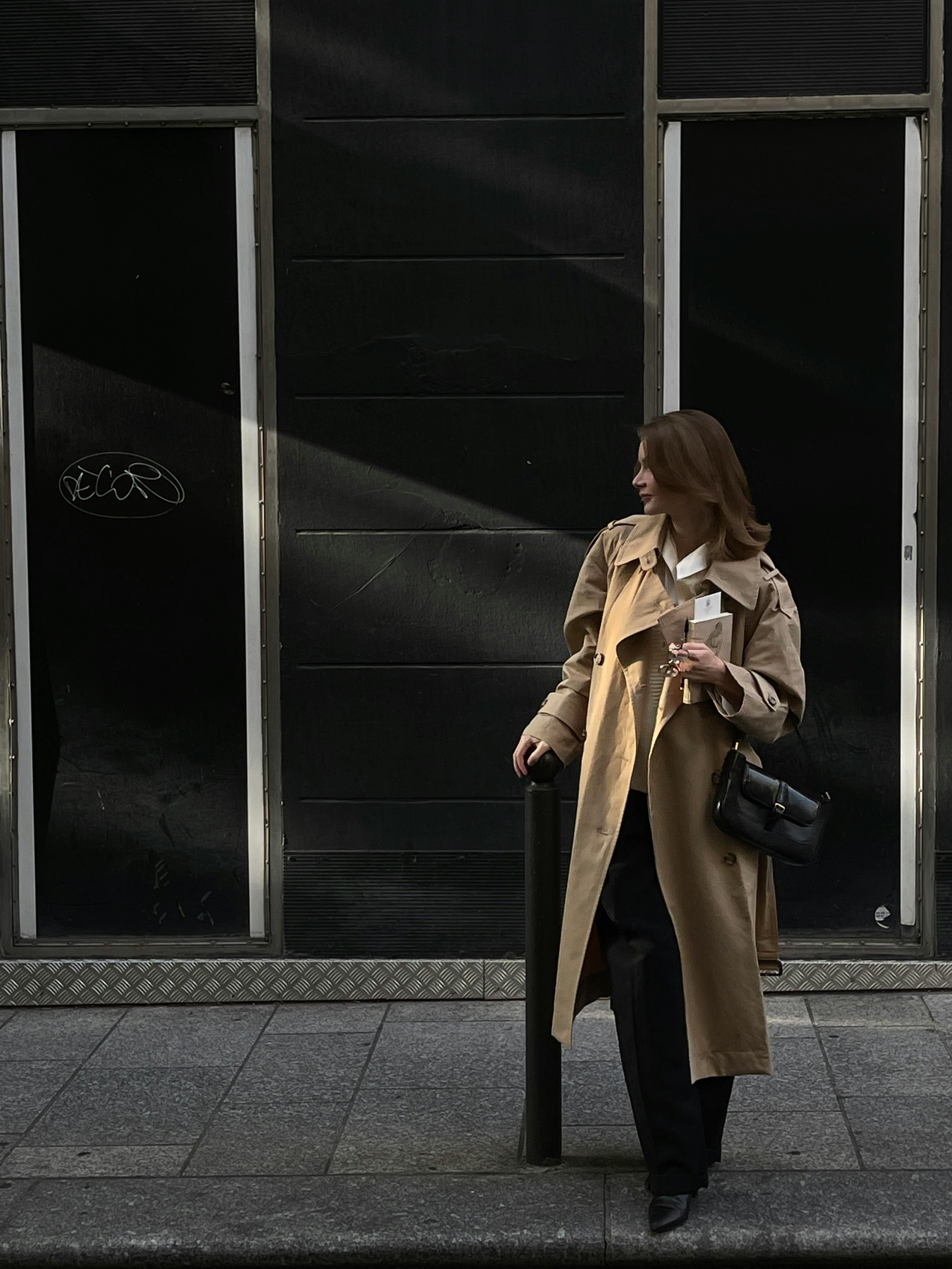 A stylish woman in a trench coat stands on a city sidewalk, exuding urban elegance.