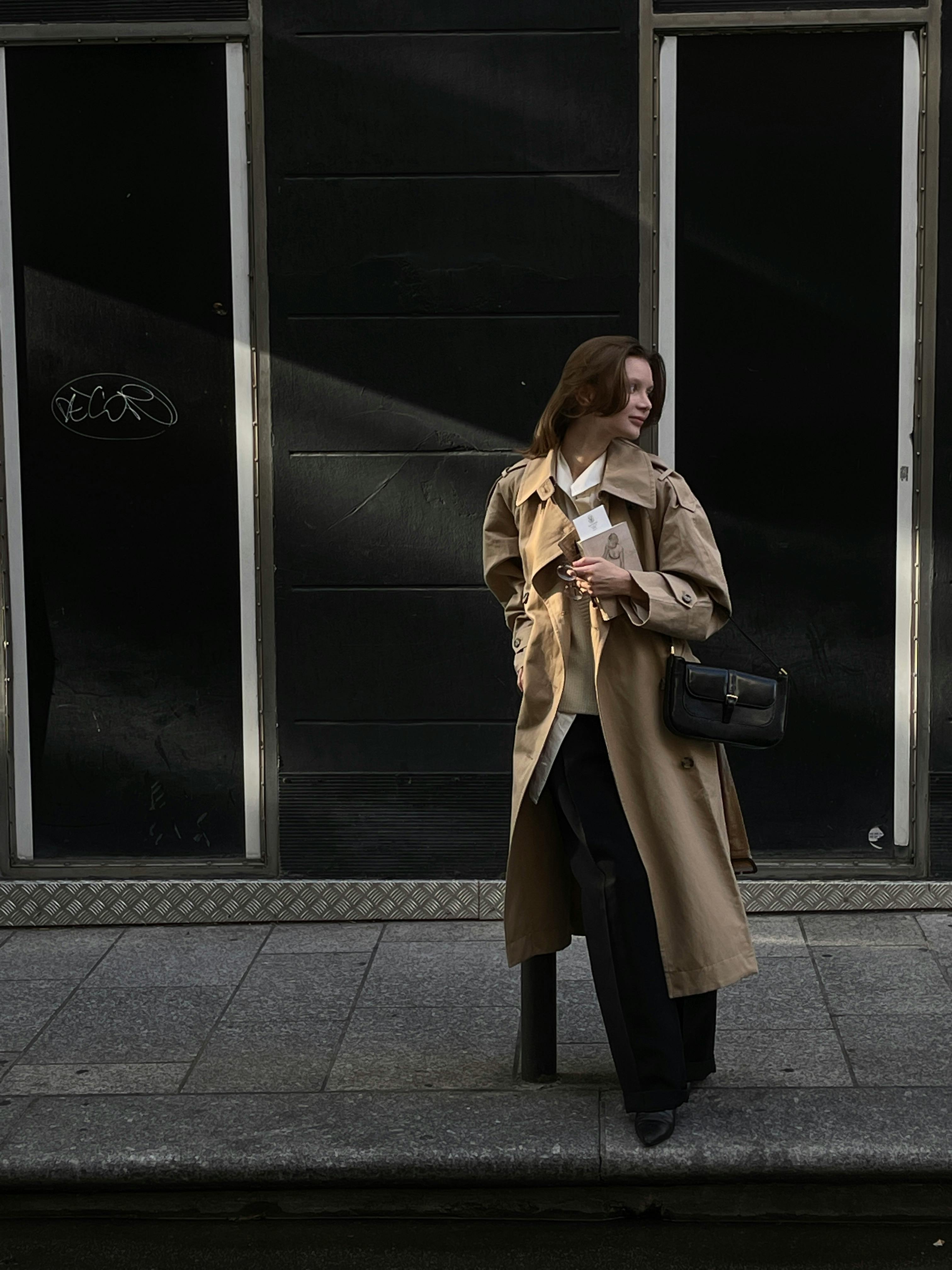 Fashionable woman in a trench coat posing on a city sidewalk, exuding modern style.