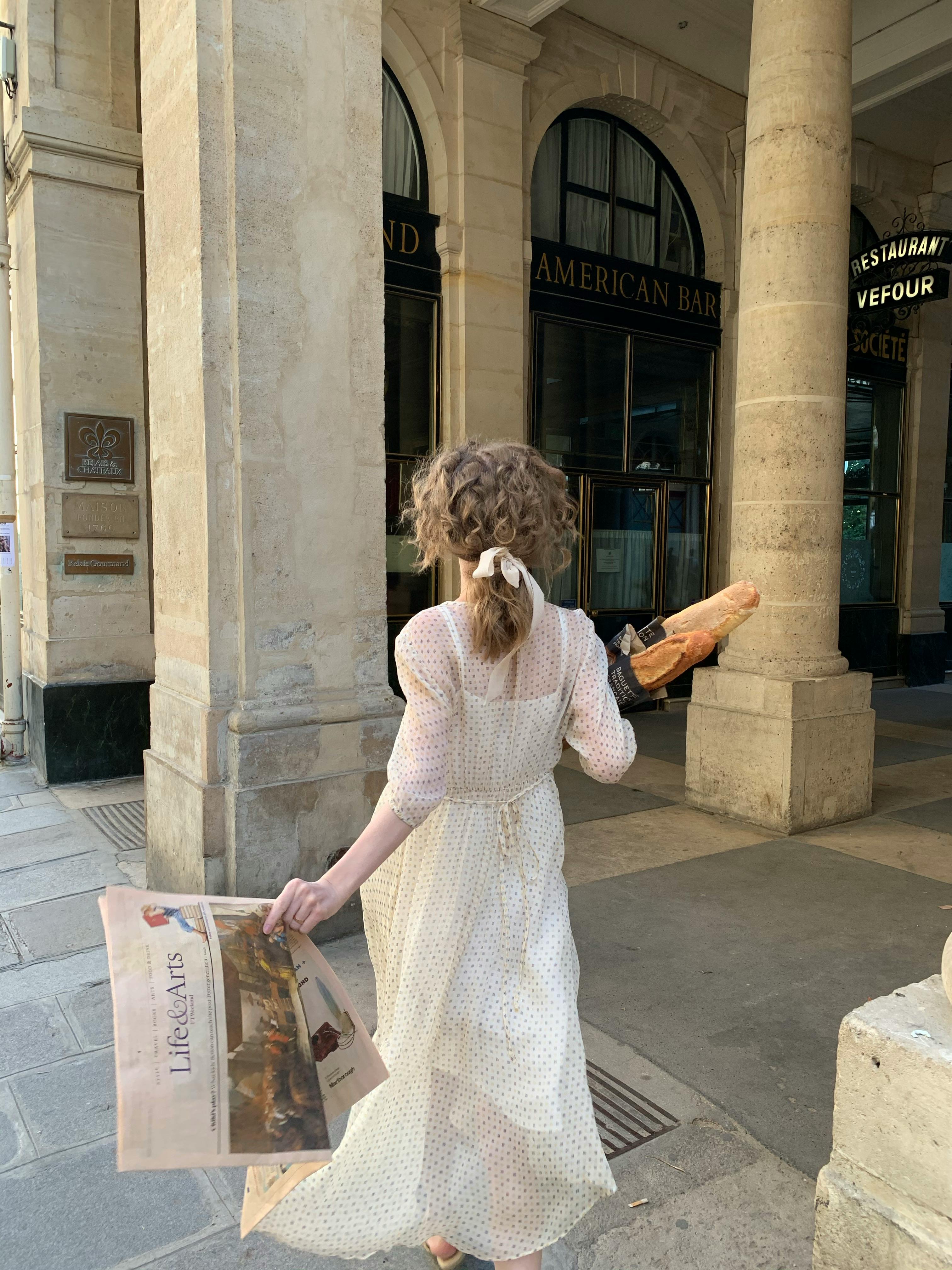 Woman in vintage dress holding newspaper and baguette, walking in Paris.