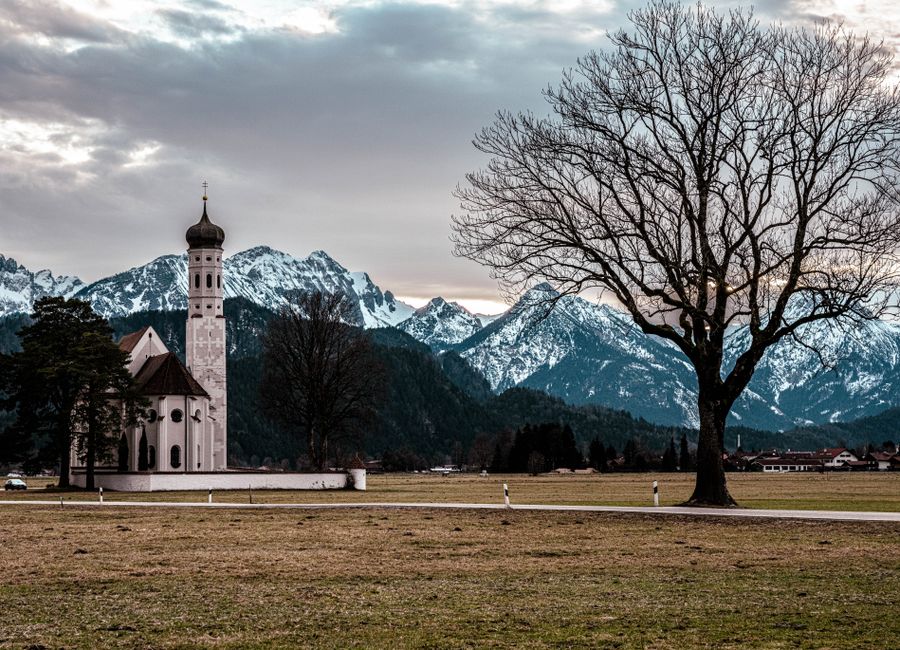 St. Coloman Church near snowy Bavarian Alps in Schwangau, Germany, under a cloudy sky.