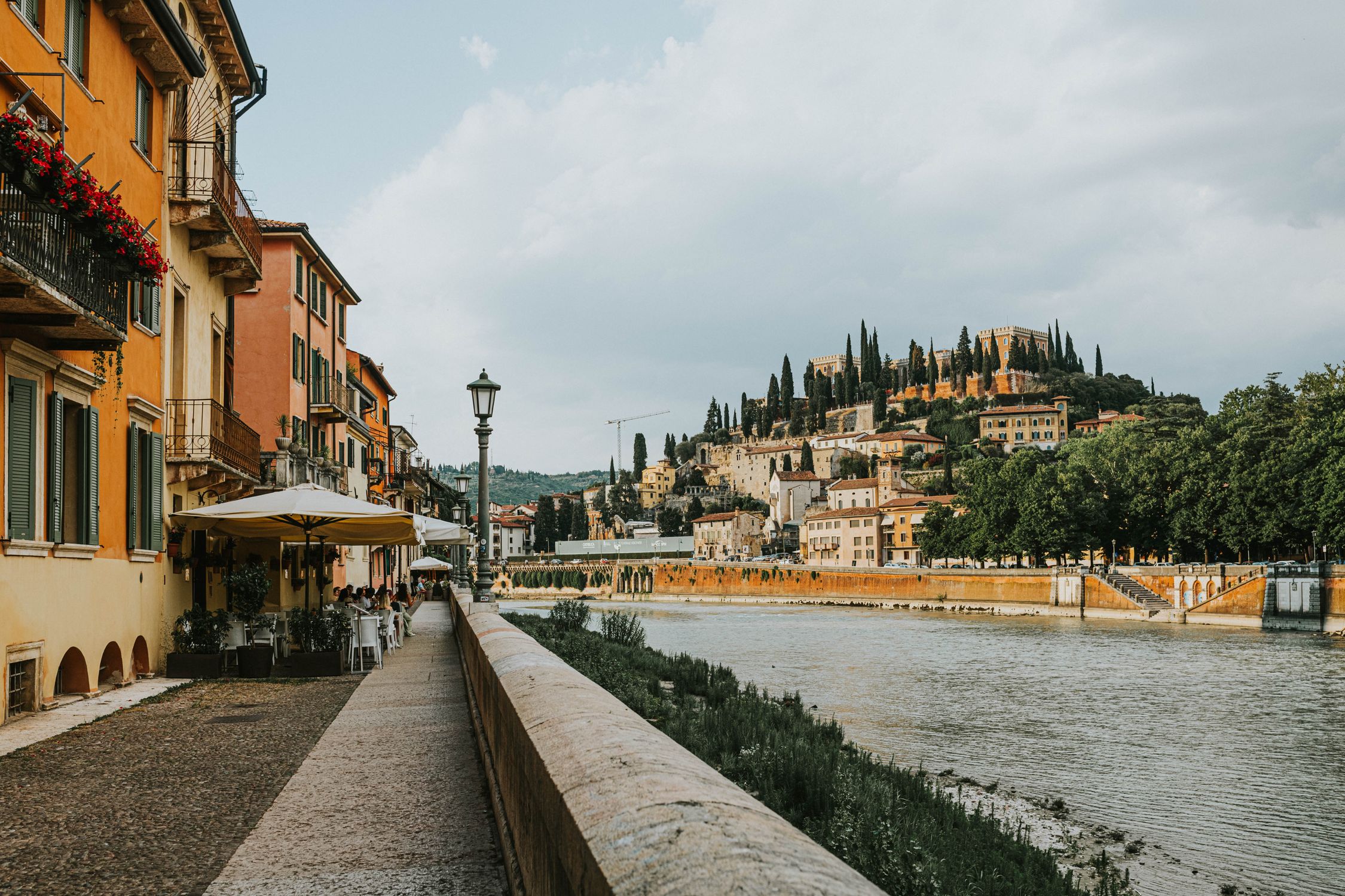 Scenic view of a riverside promenade in Verona with colorful buildings and lush greenery.