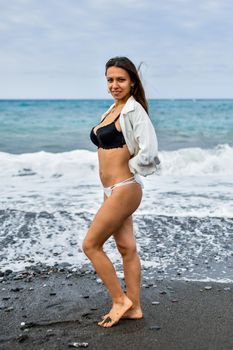 Confident woman in swimwear posing on the beach with ocean waves in the background.