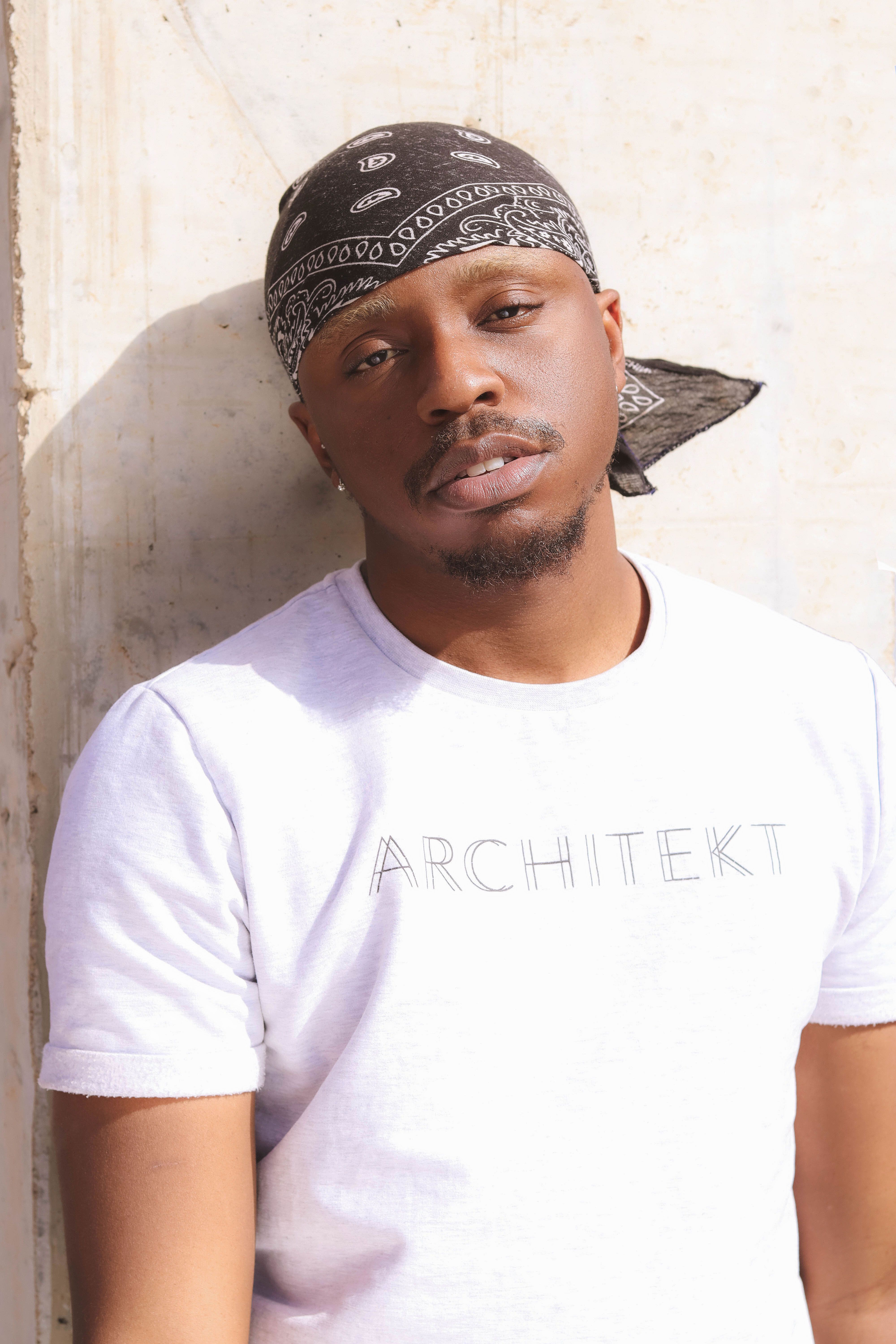 A fashionable outdoor portrait of a man leaning on a wall wearing a bandana and t-shirt.