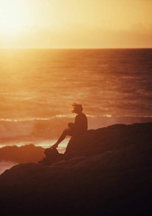 Silhouette of a Woman Sitting on a Rock on the Shore at Sunset 