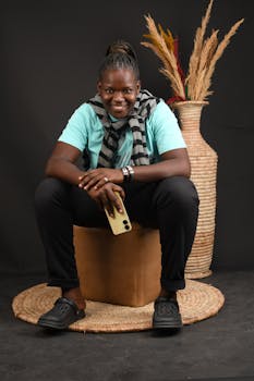 A woman dressed casually is seated with a smartphone, smiling, against a dark background, showcasing fashion and style.