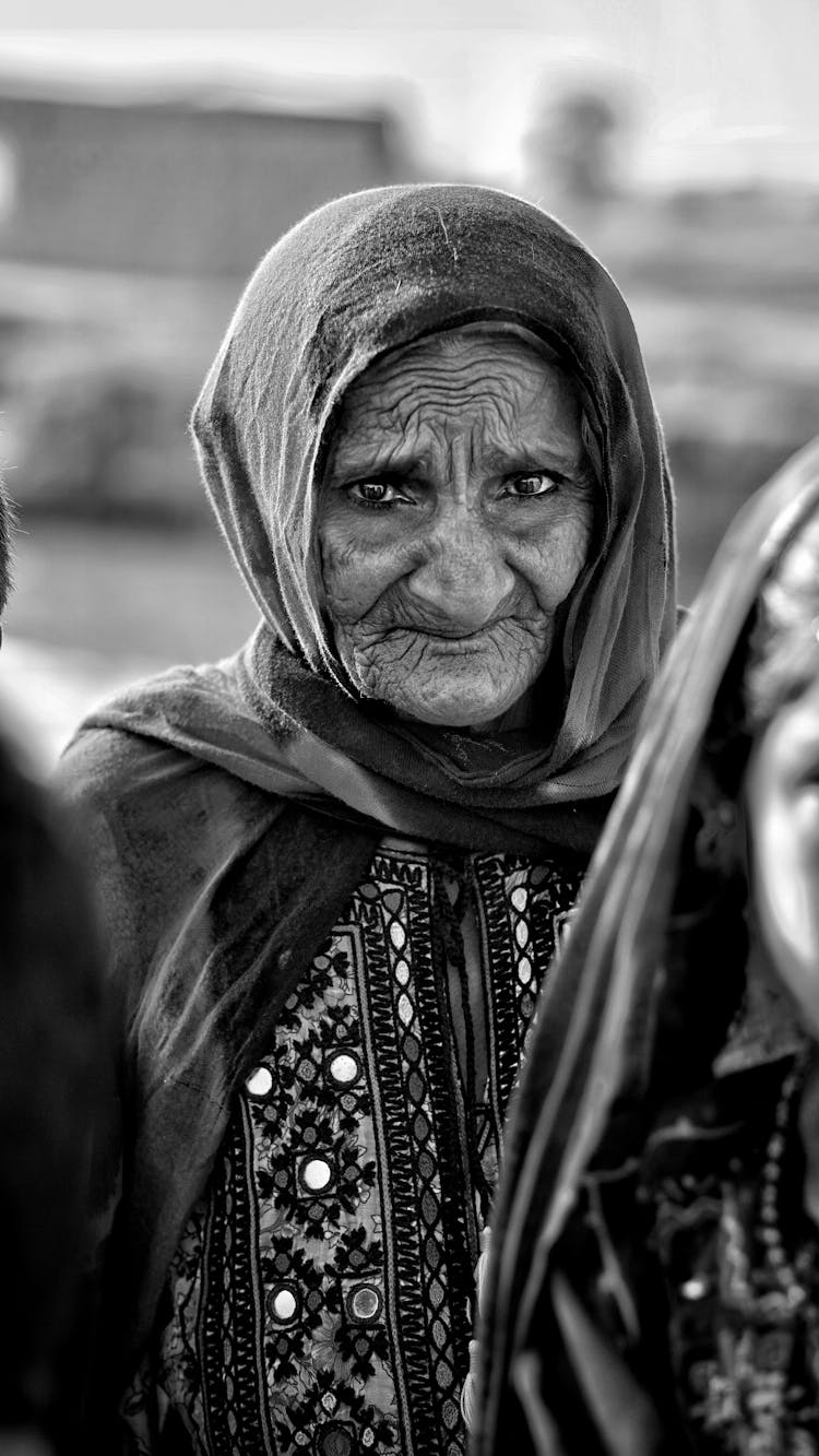 Black And White Photo Of An Elderly Woman In A Headscarf Standing Outside 