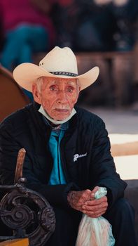 An elderly man wearing a cowboy hat sits on a city street bench holding a bag.