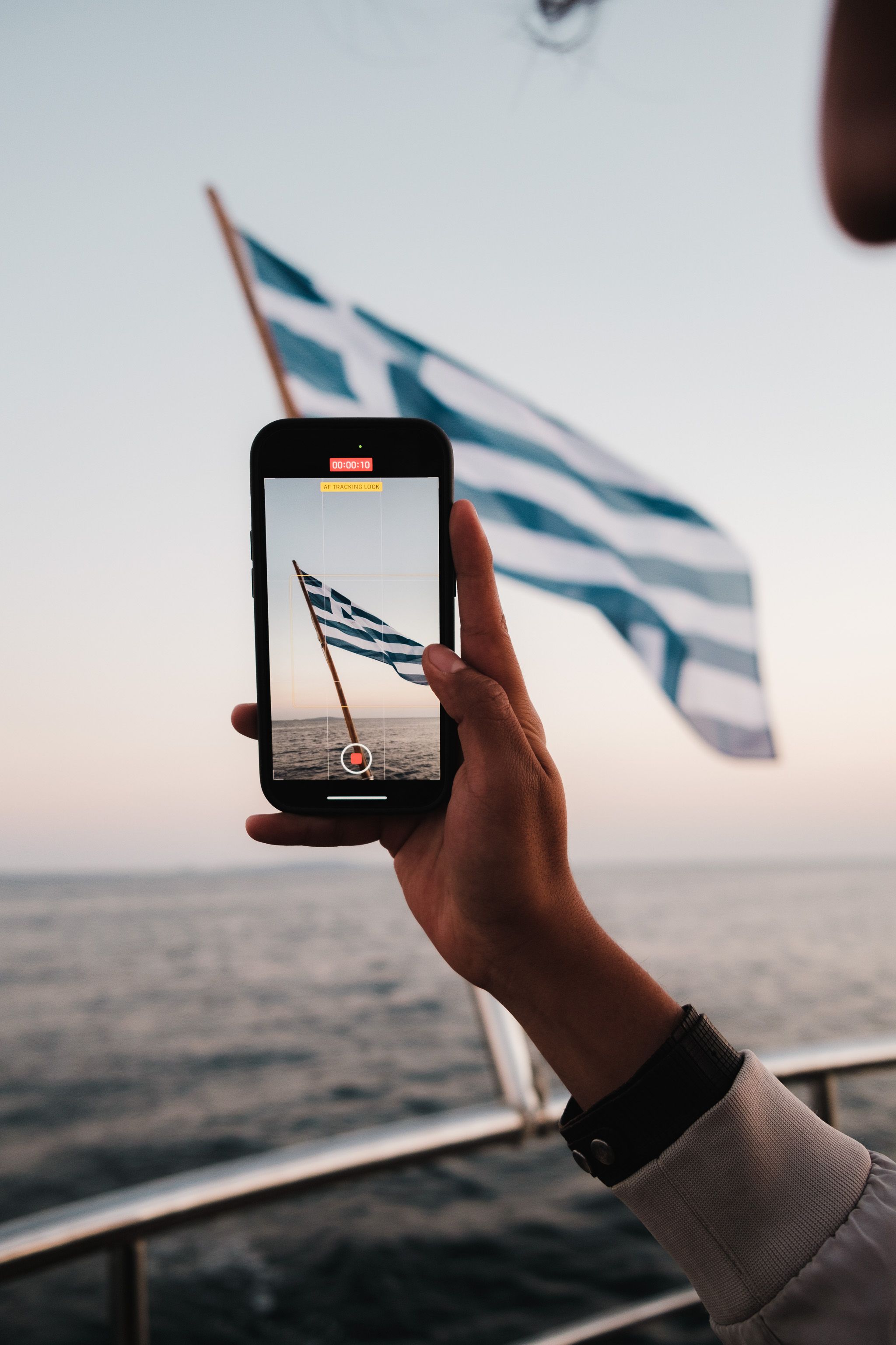 A person records the Greek flag on a smartphone while sailing during a serene day.