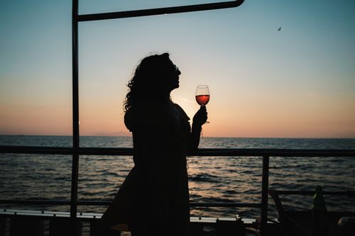 Free A woman silhouetted against a sunset over the ocean, enjoying a glass of wine on a sailboat. Stock Photo