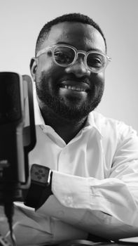 Portrait of a smiling man wearing glasses and shirt, sitting by a microphone, in black and white.