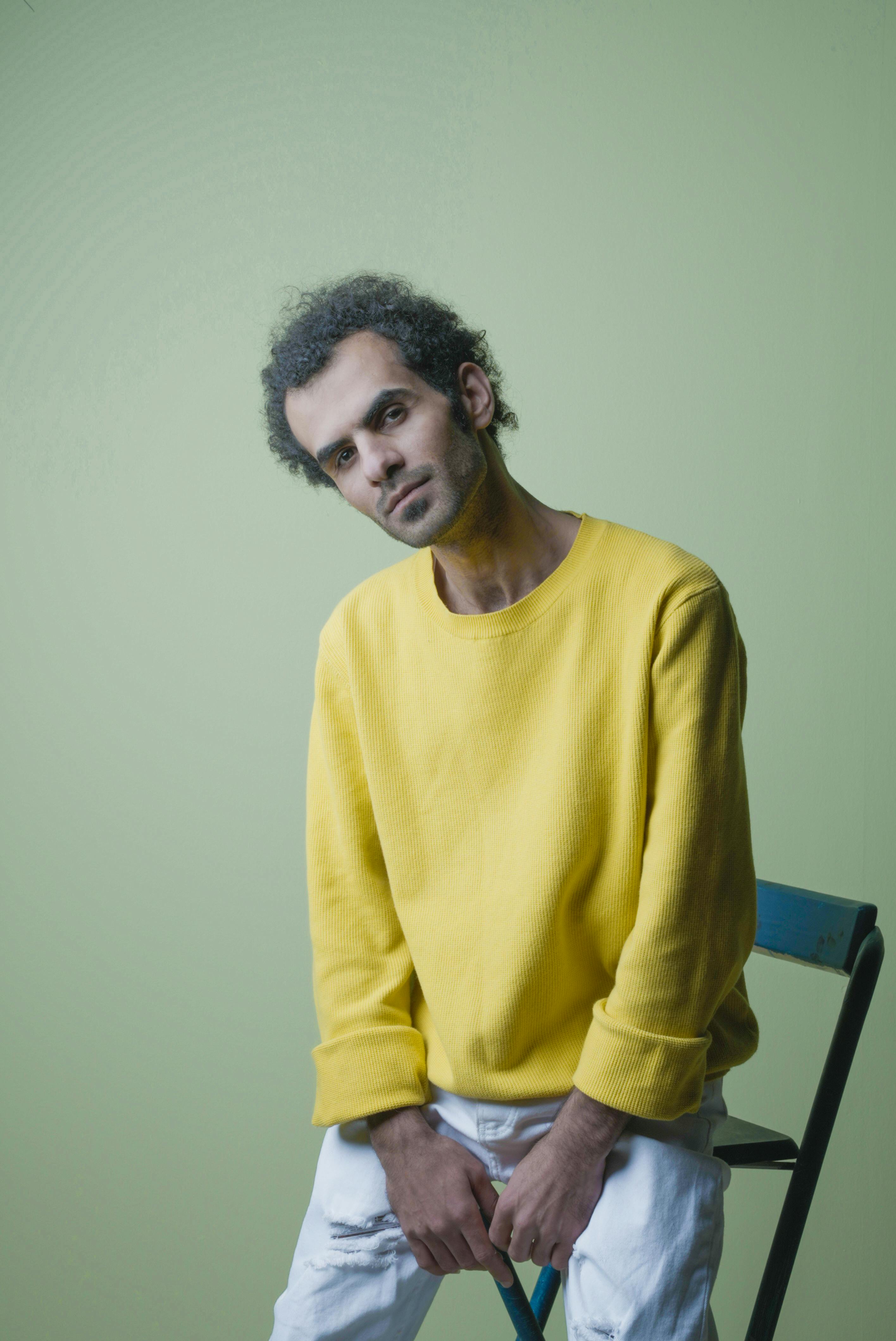 Man wearing yellow sweater sitting on a chair against a green background.