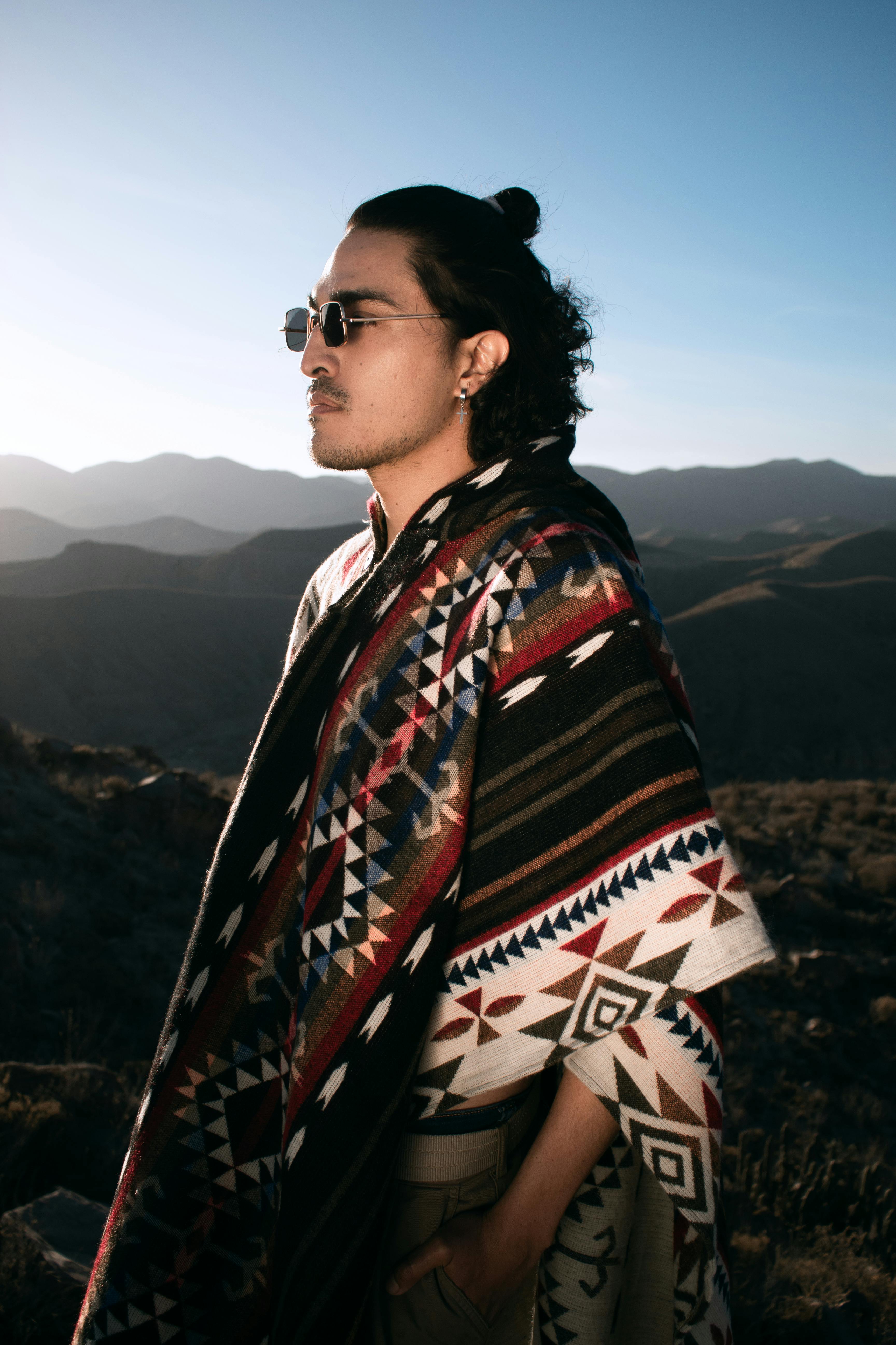 A man wearing a colorful poncho and sunglasses poses outdoors with mountains in the background.