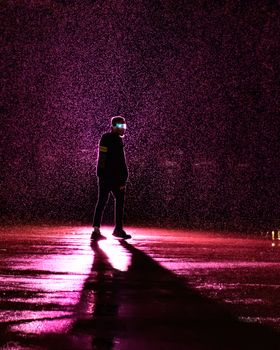 A lone figure stands under vibrant pink rain, casting a dramatic, elongated shadow on the wet ground.