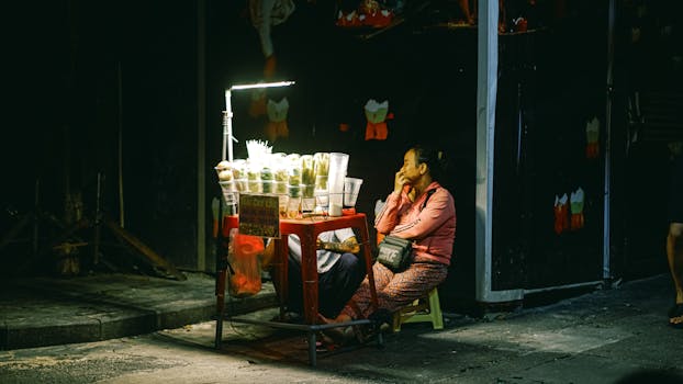 Asian woman sits at a night market stall selling street food under a bright lamp.