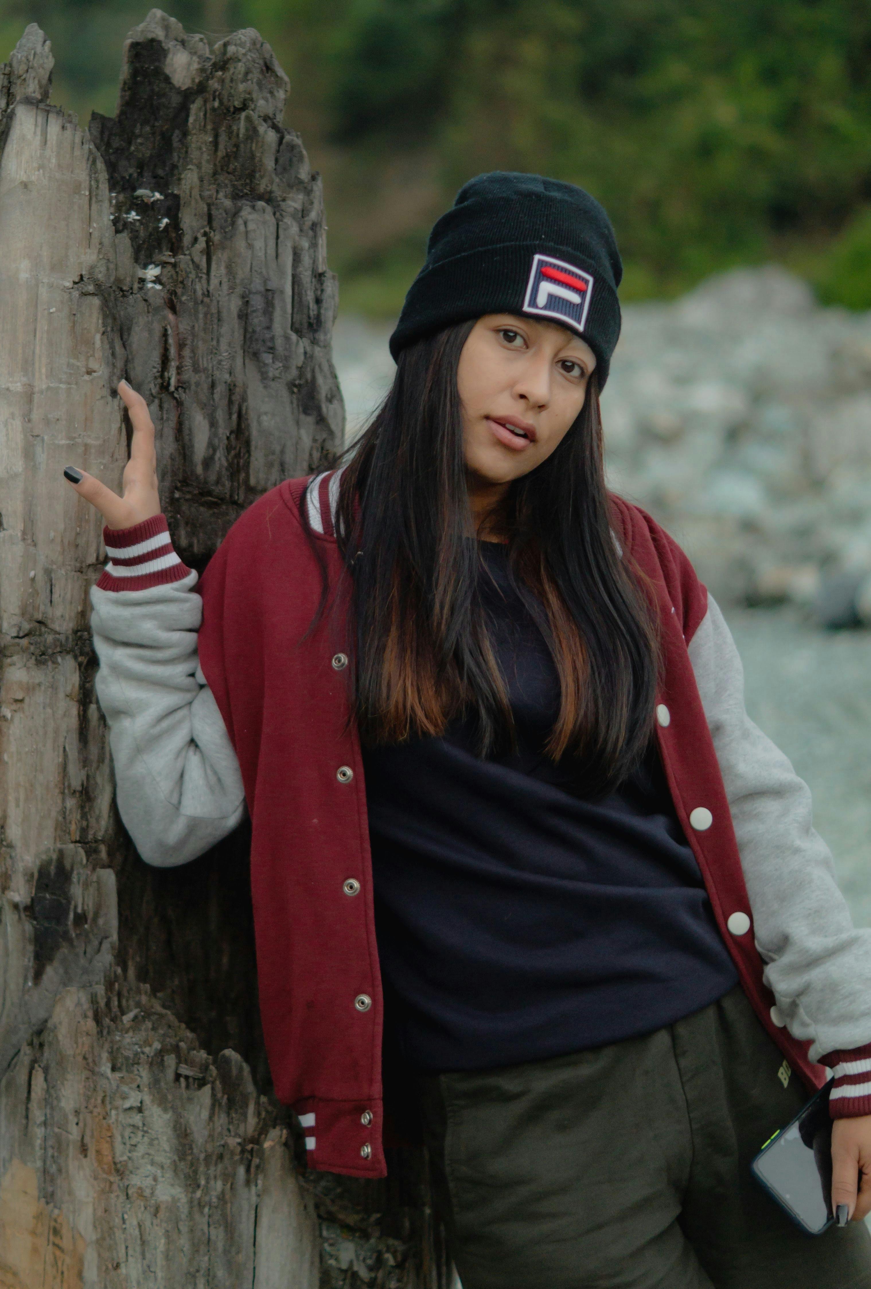 Casual photo of a young woman posing by a tree trunk outdoors, wearing a jacket and beanie.