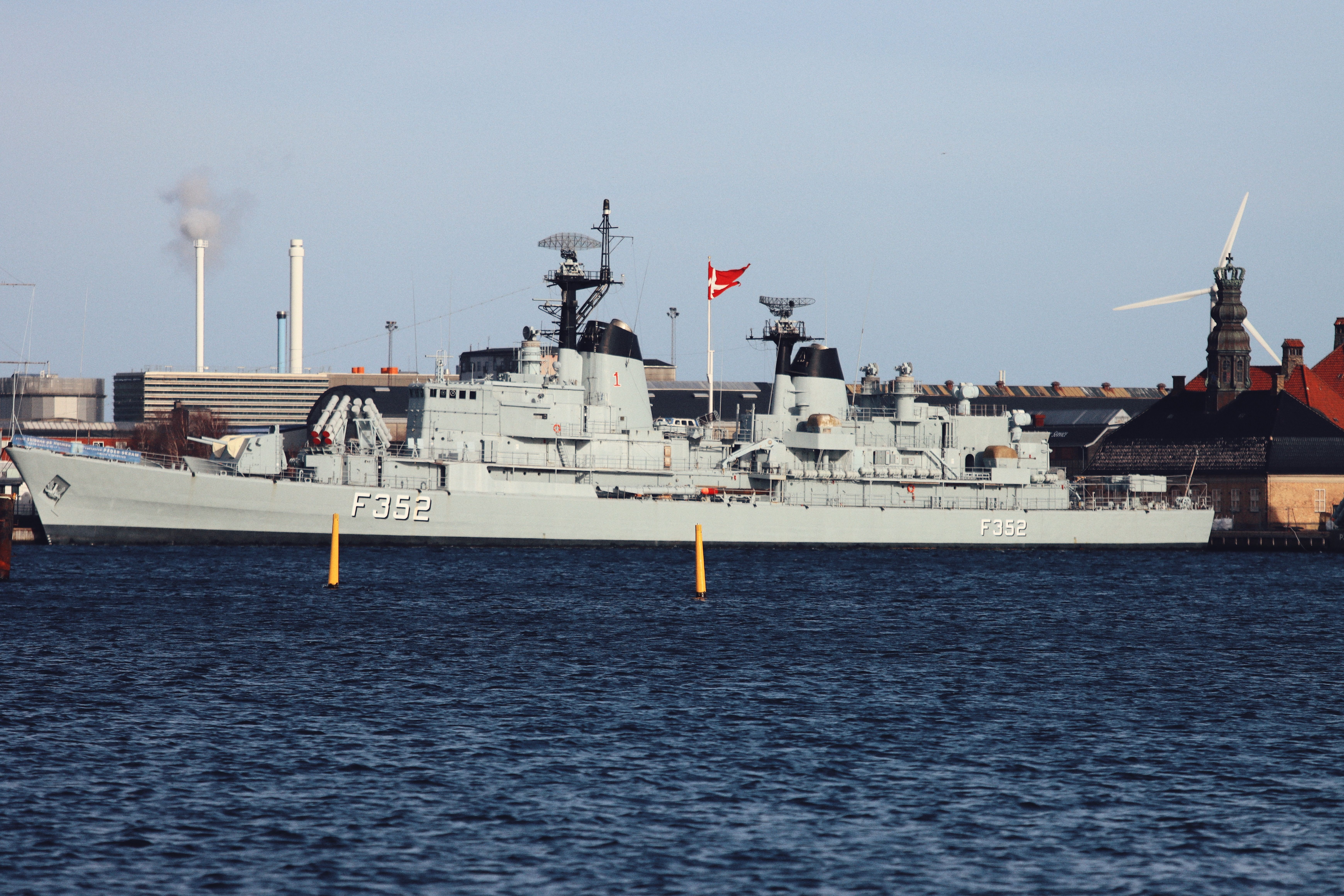 Navy ship F352 moored in a harbor with clear sky and industrial backdrop.