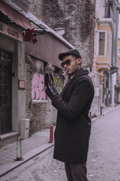 Free A fashionable young man wearing sunglasses and gloves, smoking in an urban alleyway. Stock Photo