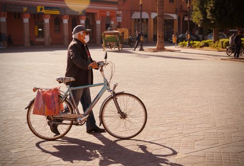 Free Elderly man wearing a mask walking a bicycle on a sunny day in a town square. Stock Photo