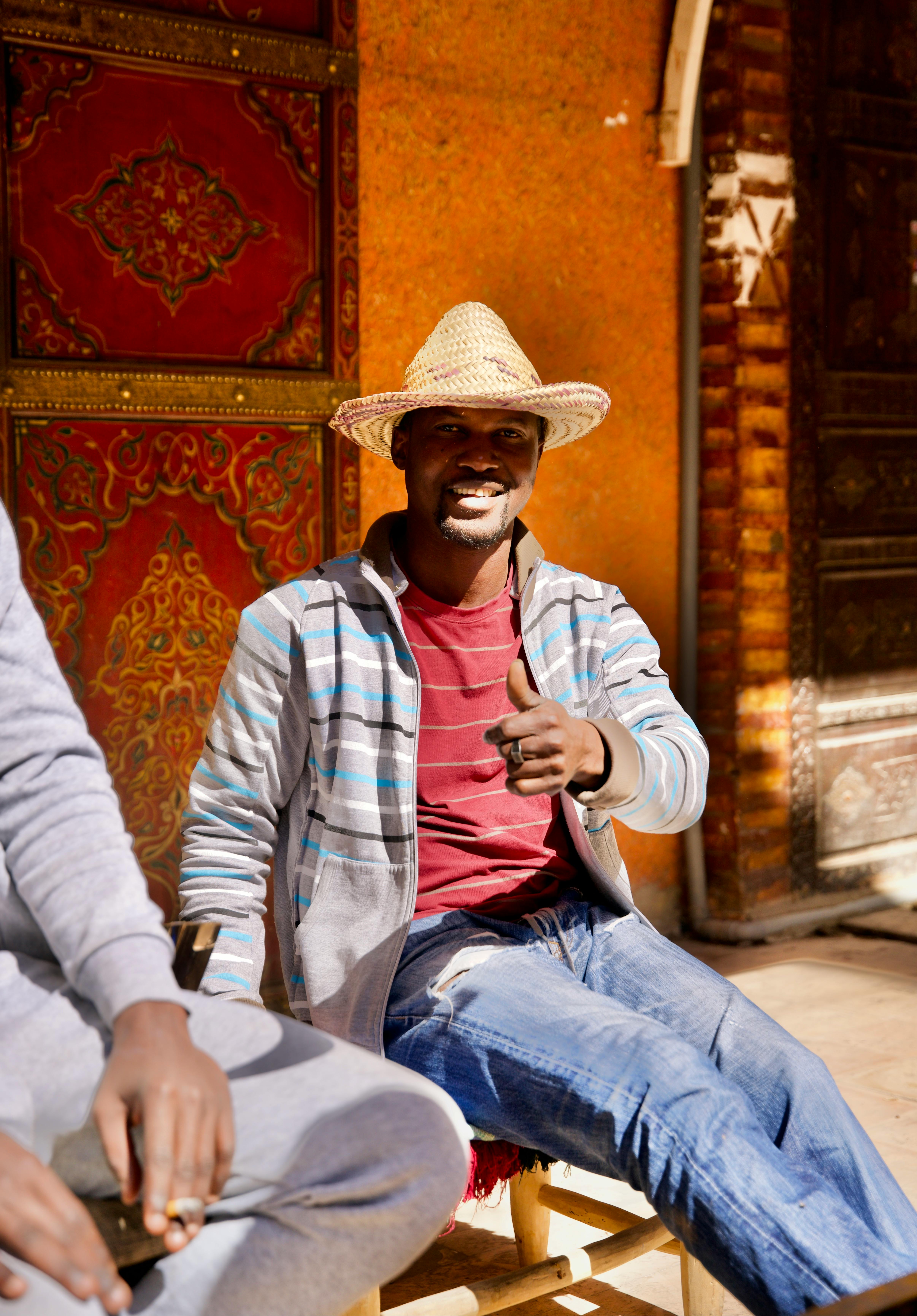 A cheerful man in casual attire and hat gives a thumbs up while sitting in an outdoor setting.