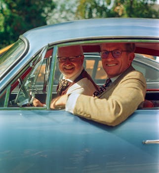 Two men in eyeglasses smiling inside a vintage blue car on a sunny day.