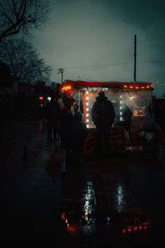 A silhouette of people gathered around a lit street food stall during a rainy night.