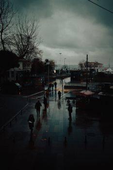 A dramatic urban street scene on a rainy day with people walking and reflections on wet pavement.