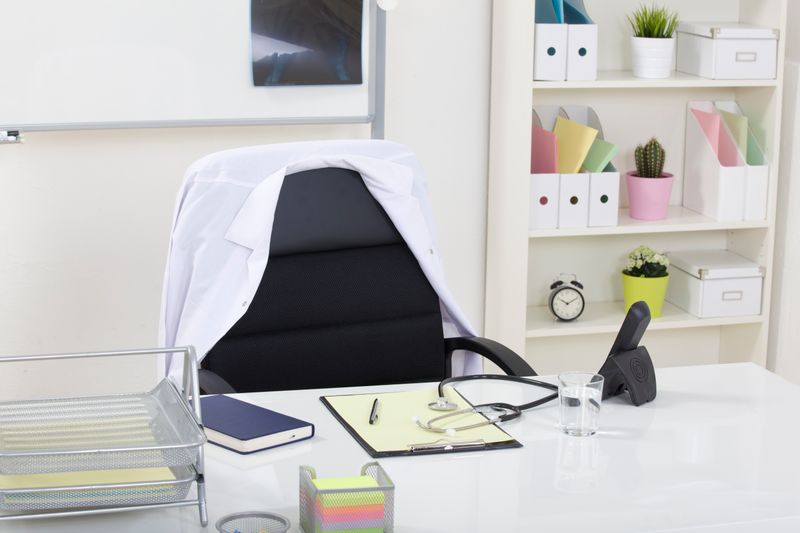 A clinician's desk viewed from the patient seat: white doctor's coat draped on the chair, a stethoscope coiled across a yellow legal pad, a whiteboard with an X-ray clipped on the back wall.