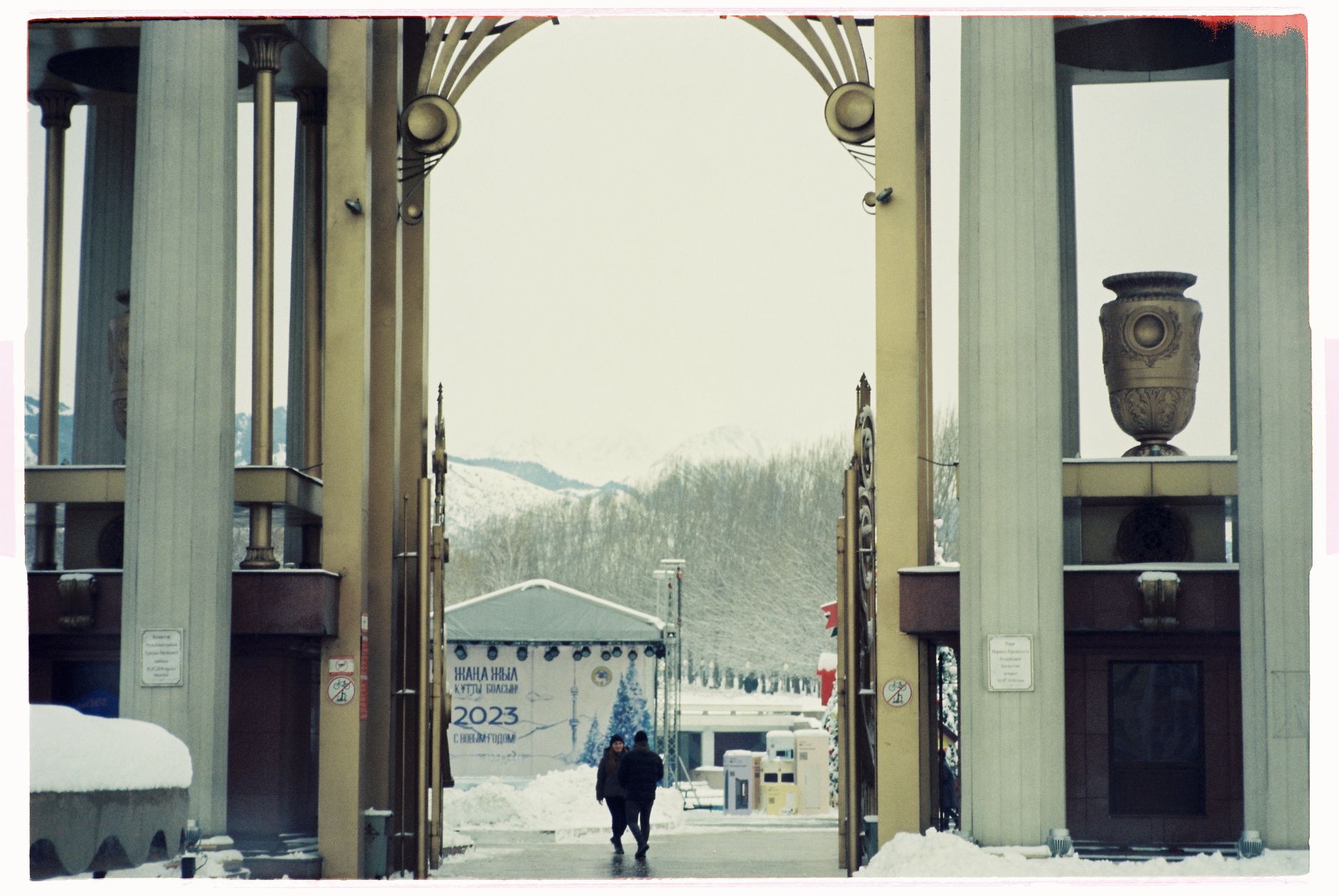 People walk through an ornate gate with winter snow in the background.