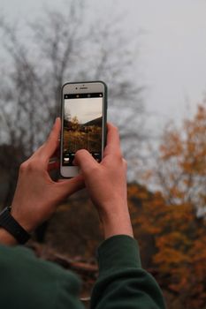 A person in a green jacket taking a photo of autumn trees using a smartphone.