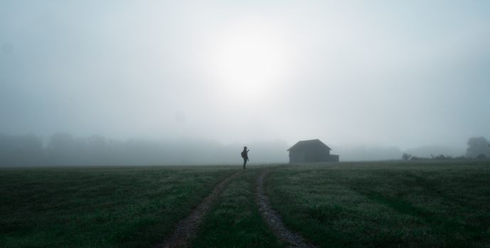 A lone figure on a dirt road through a misty field, with a barn in sight.