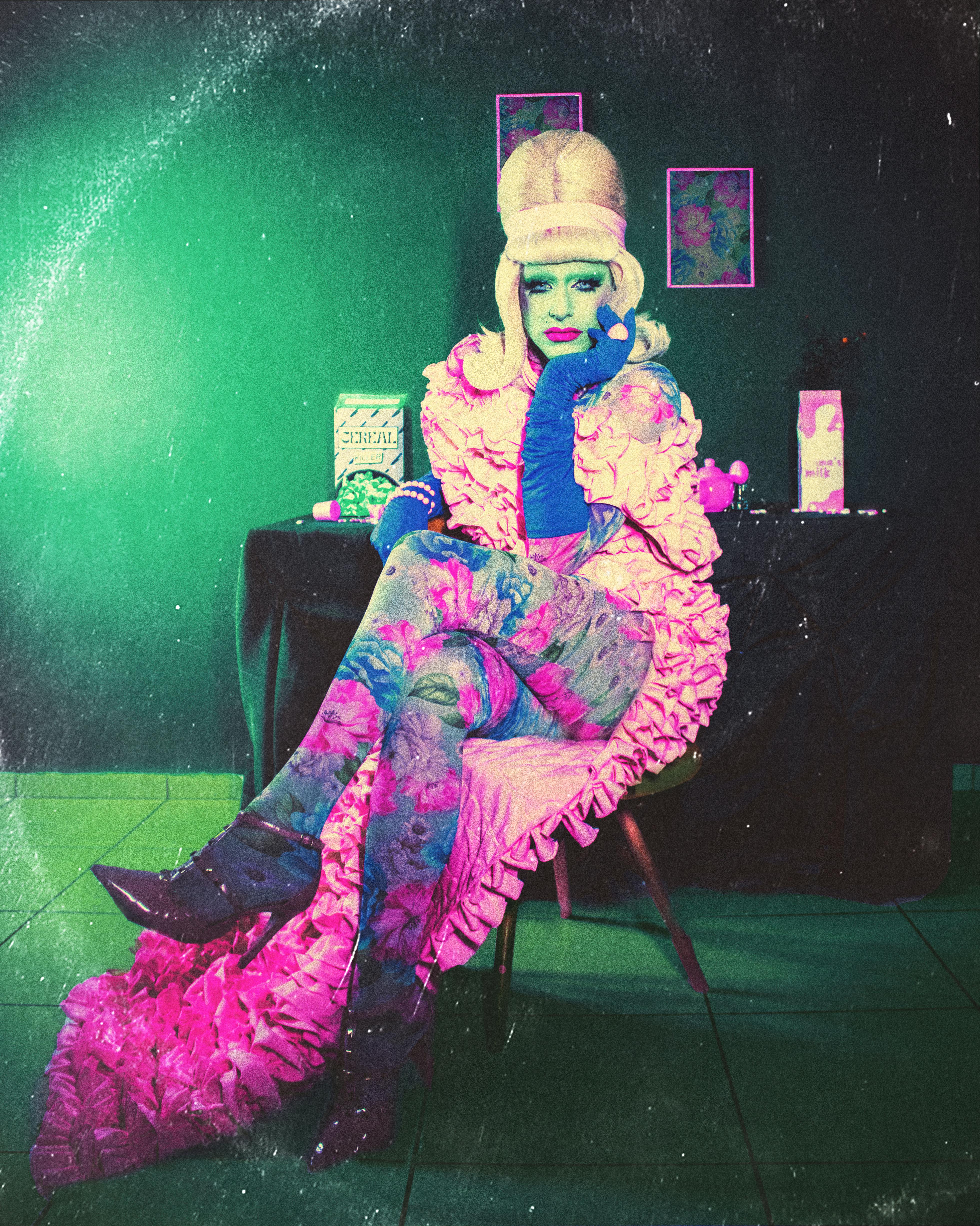 A colorful drag queen in unique fashion with retro tones sitting stylishly indoors.