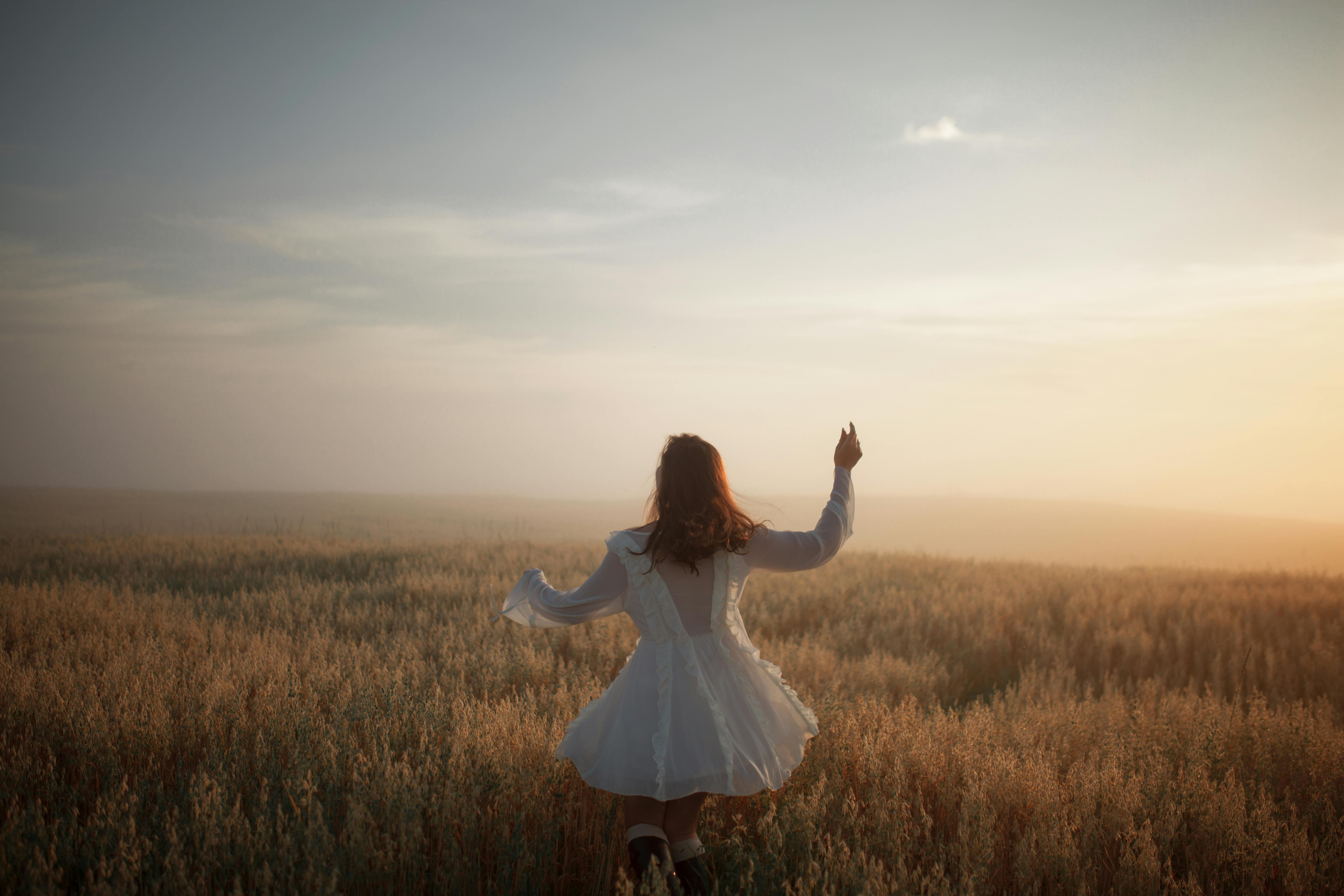 Back view of woman in white dress dancing in a sunlit rural field at sunset.