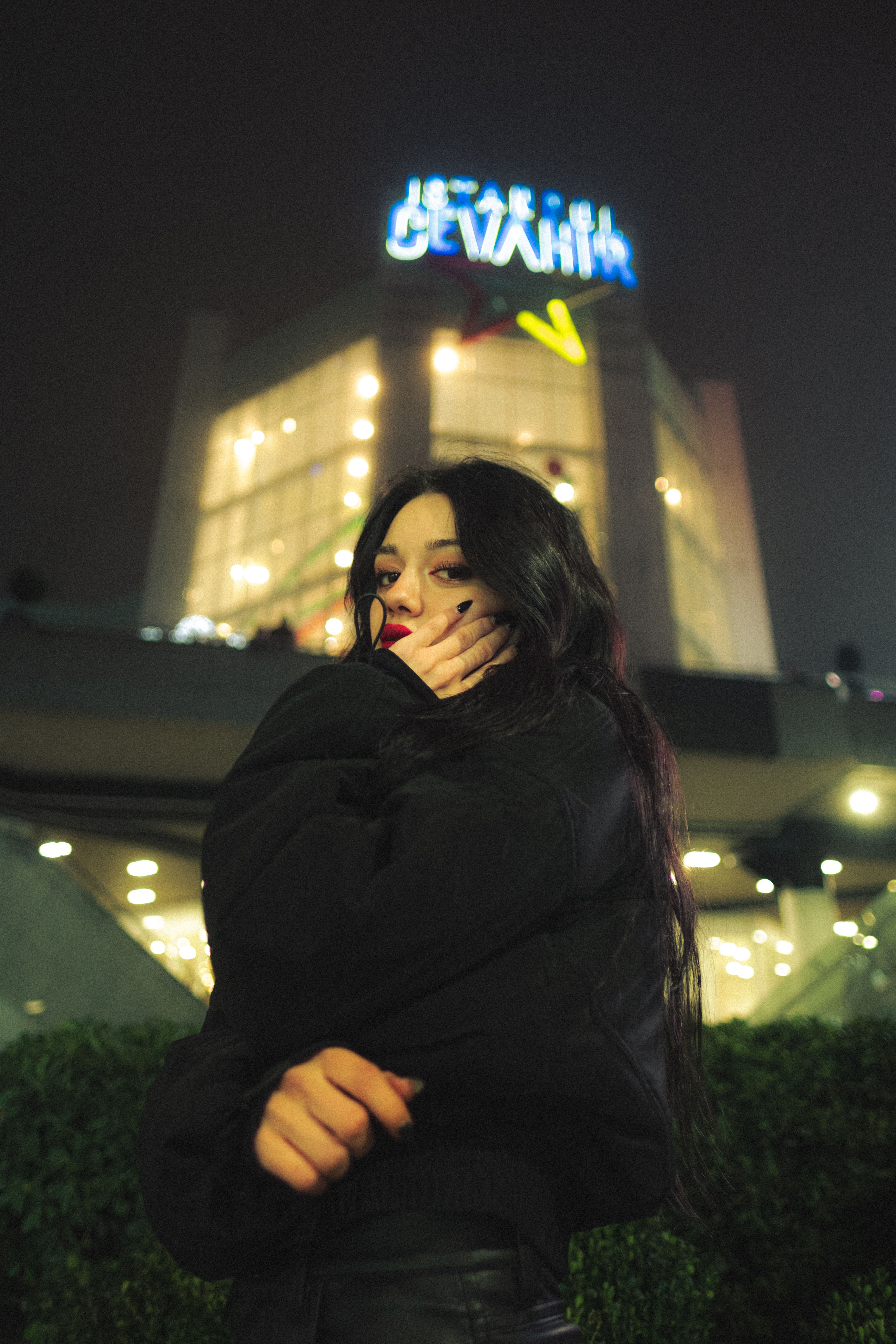 Moody nighttime portrait of a woman in front of an illuminated urban building.